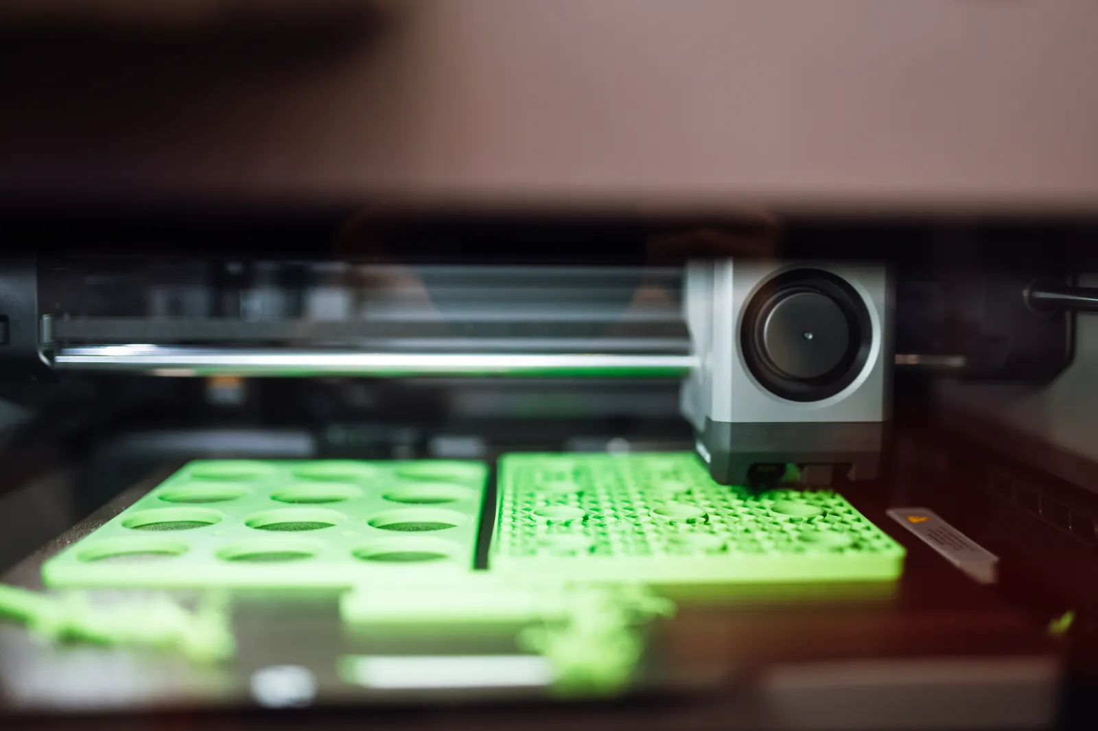 A desktop 3D printer laying down green plastic parts on its print bed, with the print head in motion