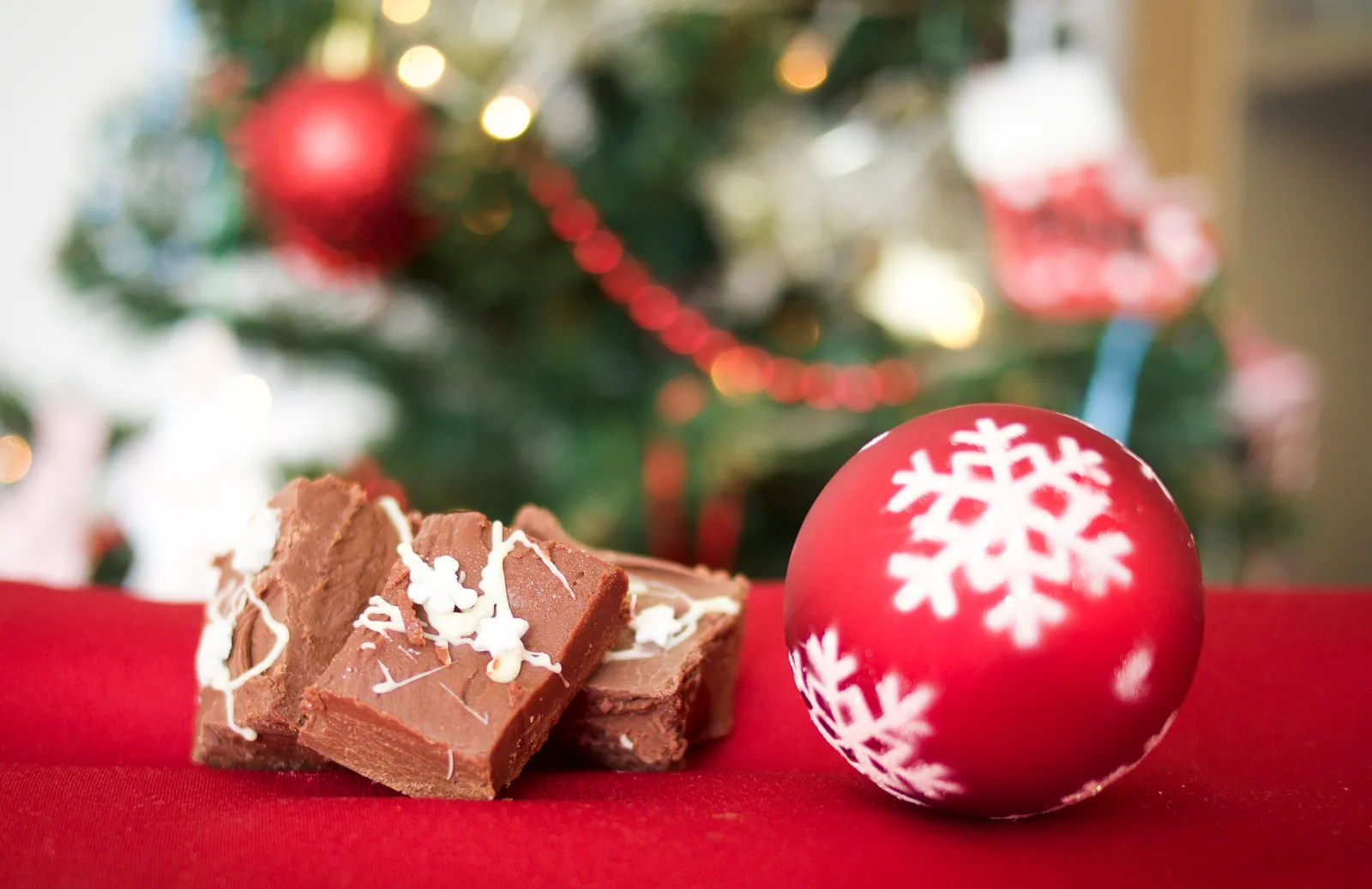 Squares of chocolate fudge and a red snowflake bauble on a red tablecloth in front of a lit Christmas tree
