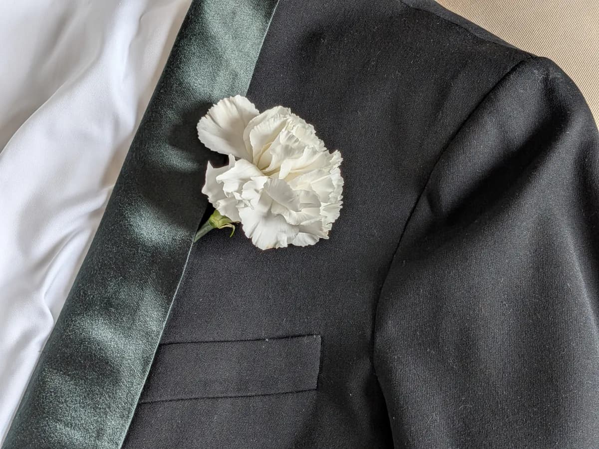A white carnation boutonniere pinned to the lapel of a black tuxedo jacket with a satin shawl collar