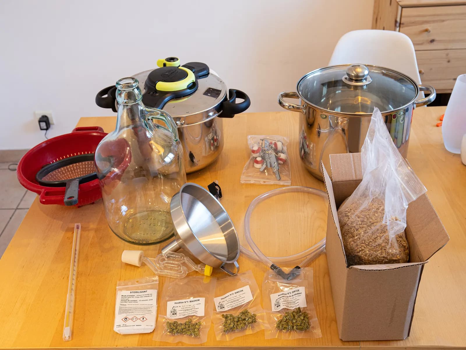 A beginner homebrewing setup laid out on a wooden table: glass jug, two stockpots, funnel, tubing, airlock, thermometer, hop pellets and a box of malted grain