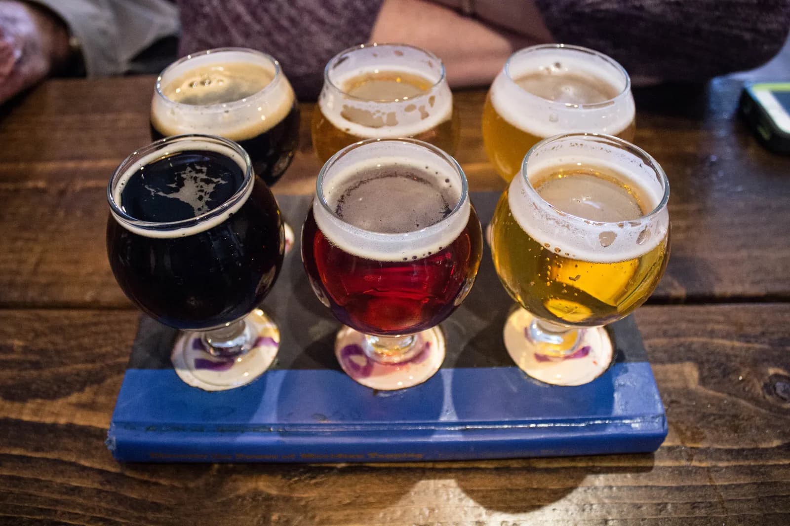 A tasting flight of six beers, from a dark stout to a golden pale ale, in small stemmed glasses on a blue board on a wooden table