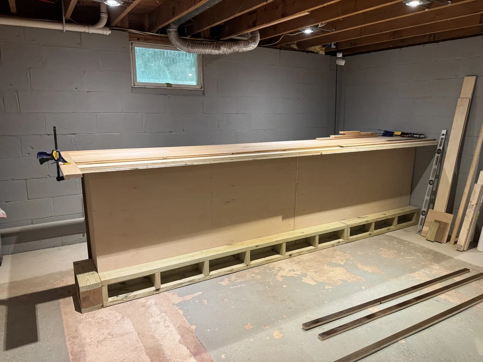 A long wooden bar counter being built in an unfinished basement, with clamps on the top and spare boards leaning against a gray block wall