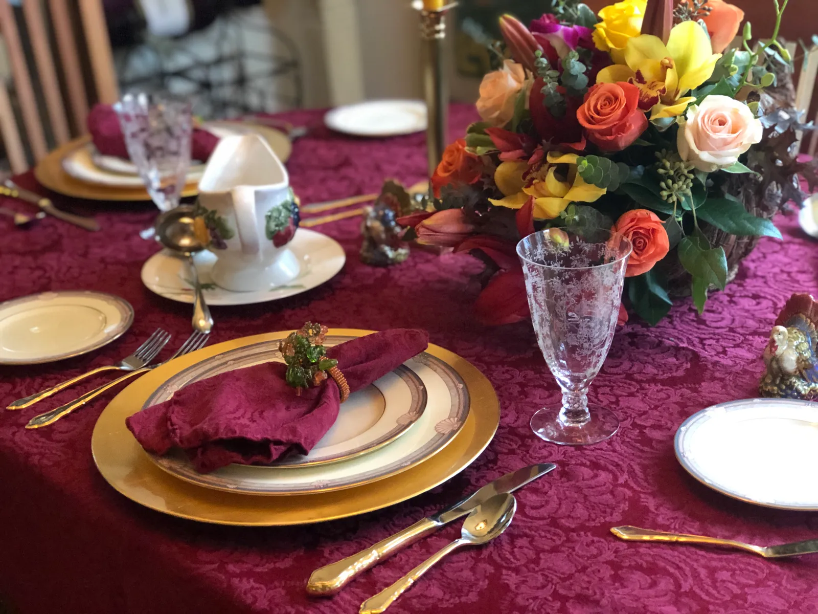 A Thanksgiving place setting on a burgundy tablecloth, with a gold charger plate, a folded napkin, an etched water glass and a bouquet of orange roses and lilies
