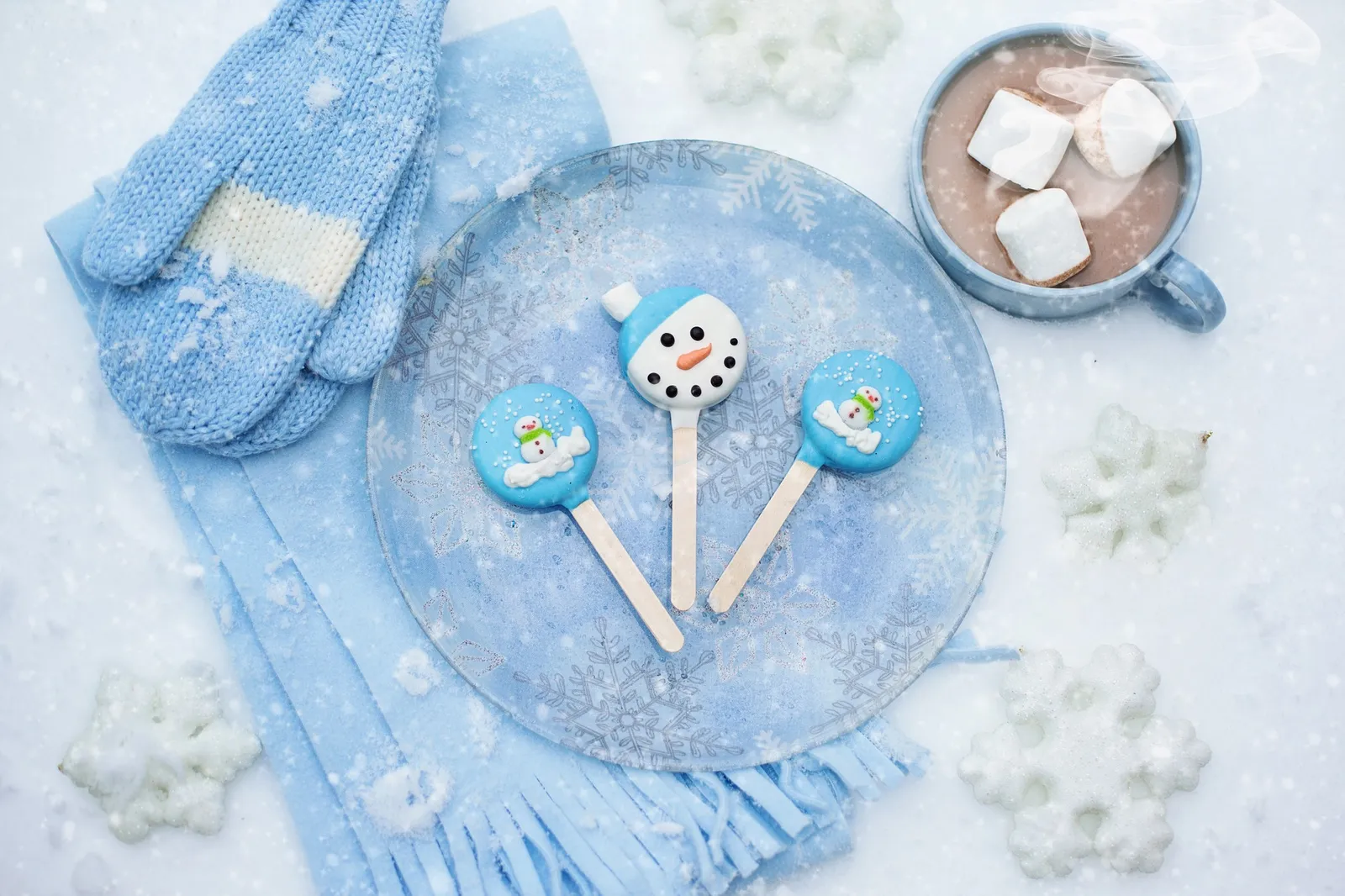 Hot chocolate with marshmallows next to blue knitted mittens, a scarf and snowman cake pops on a frosted plate