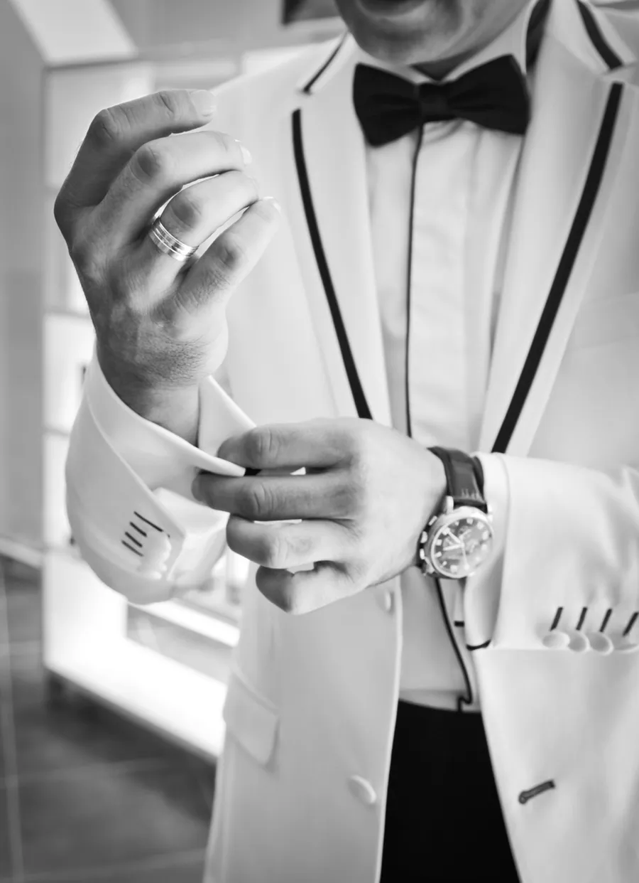 A man in a white tuxedo jacket and black bow tie adjusting his shirt cuff, wedding ring on his hand