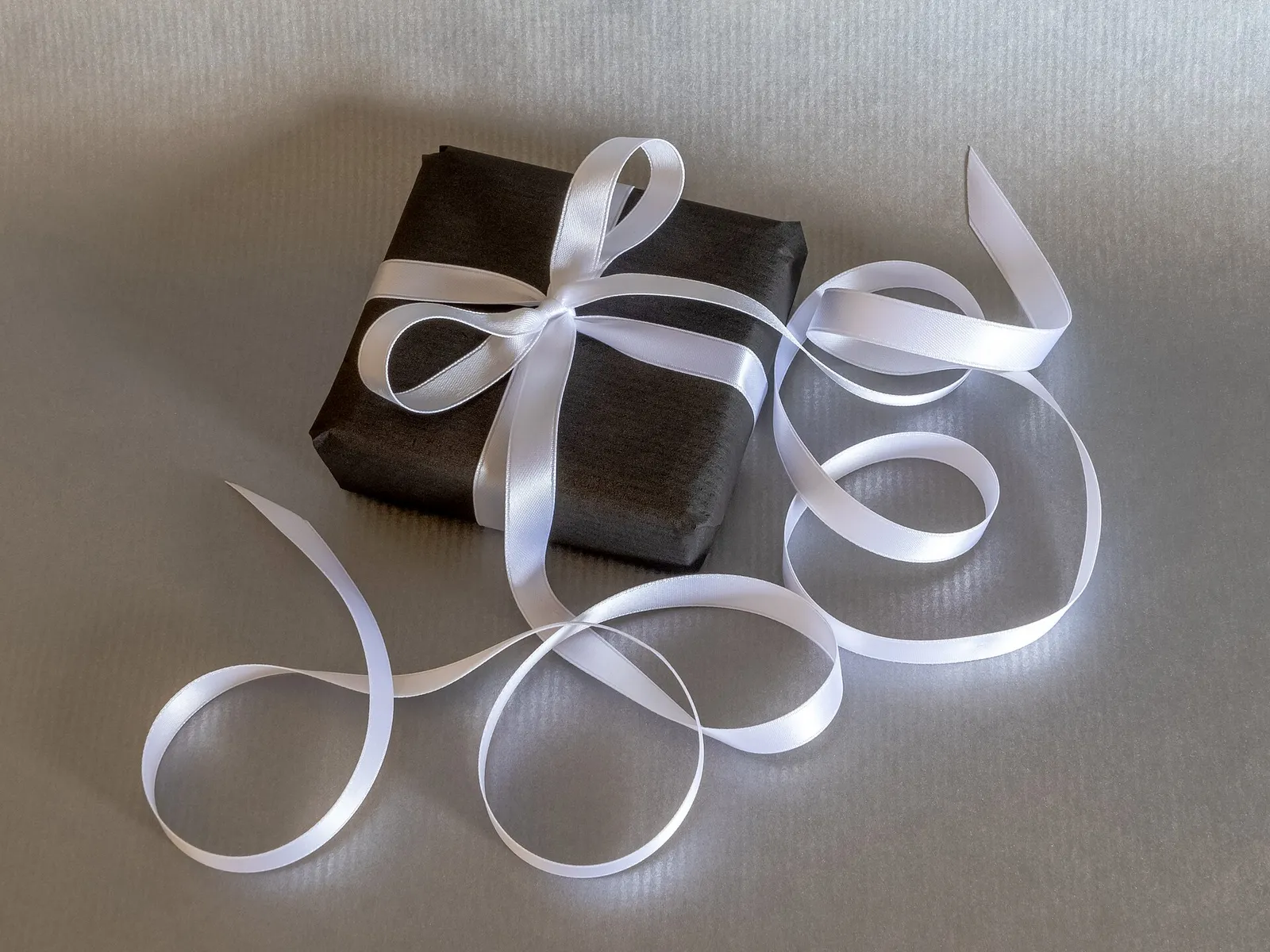 A small gift wrapped in dark paper and tied with a long white satin ribbon, on a grey surface