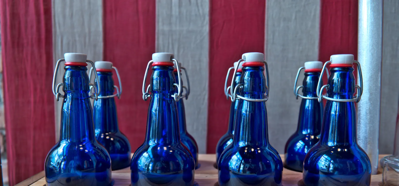 Rows of blue glass swing-top bottles with white ceramic stoppers and wire clasps, set in a wooden crate in front of a red and white striped backdrop