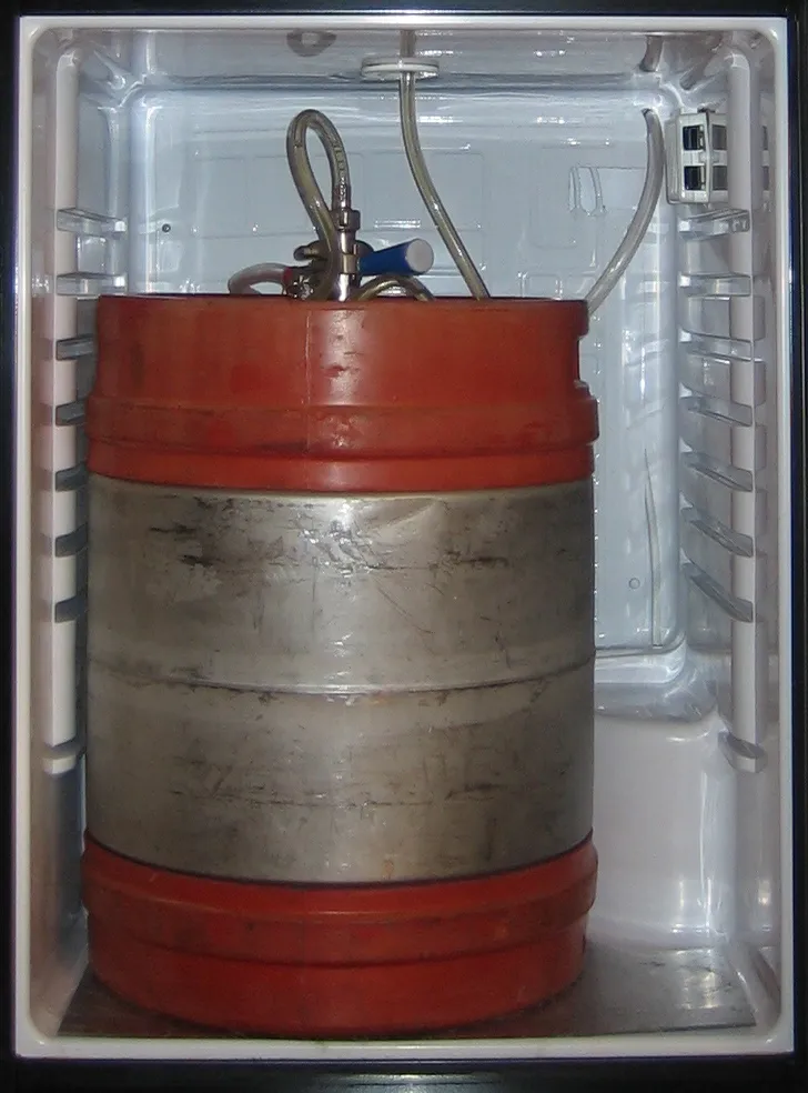 A metal beer keg with orange rubber rings standing inside a small open refrigerator fitted with a dispensing line